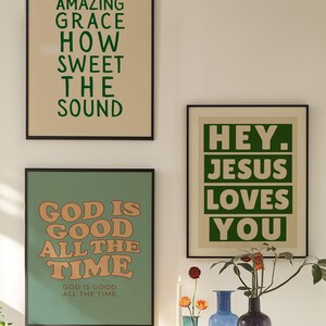Op de afbeelding: Drie ingelijste kunstdrukken met inspirerende citaten. De bovenste print luidt "AMAZING GRACE HOW SWEET THE SOUND" in groen. De middelste print zegt "GOD IS GOOD ALL THE TIME" in oranje. De derde print zegt "HEY. JESUS LOVES YOU" in groen.