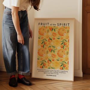 May include: A framed print with a yellow background and a repeating pattern of orange slices, white flowers, and green leaves. The print has the text "FRUIT of the SPIRIT" at the top and a Bible verse from Galatians 5:22-23 at the bottom.