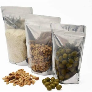 Peut inclure: Trois sachets transparents &agrave; fond plat, doubl&eacute;s d'argent, remplis d'aliments. L'un contient du riz blanc, un autre des cacahu&egrave;tes et le troisi&egrave;me des olives vertes. Un tas de cacahu&egrave;tes et un petit groupe d'olives sont devant les sachets.