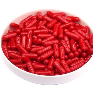 Peut inclure: Un bol blanc rempli de nombreuses g&eacute;lules rouges. Les g&eacute;lules sont de forme cylindrique avec des extr&eacute;mit&eacute;s arrondies et un aspect brillant et translucide. Le bol est pos&eacute; sur un fond blanc uni, mettant en valeur la couleur vive des g&eacute;lules.
