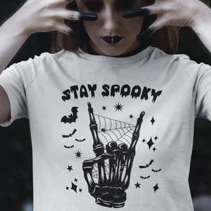 May include: White t-shirt with a black skeleton hand making a rock and roll sign. The hand is surrounded by bats, stars, and a spider web. The text "STAY SPOOKY" is above the hand.