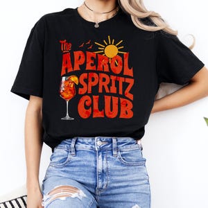 K&ouml;nnte beinhalten: Ein schwarzes T-Shirt mit einem Grafikdesign, auf dem "The Aperol Spritz Club" in roten und orangen Buchstaben steht. Die Grafik enth&auml;lt auch ein Glas Aperol Spritz, eine Sonne und V&ouml;gel.