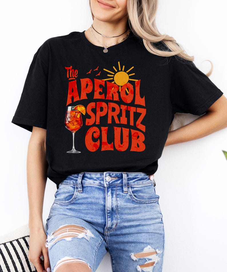 K&ouml;nnte beinhalten: Ein schwarzes T-Shirt mit einem Grafikdesign, auf dem "The Aperol Spritz Club" in roten und orangen Buchstaben steht. Die Grafik enth&auml;lt auch ein Glas Aperol Spritz, eine Sonne und V&ouml;gel.