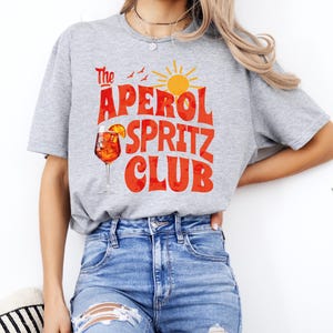 K&ouml;nnte beinhalten: Ein graues T-Shirt mit dem Text "The Aperol Spritz Club" in roten und orangen Buchstaben. Das Design enth&auml;lt auch ein Glas Aperol Spritz, eine Sonne und V&ouml;gel.