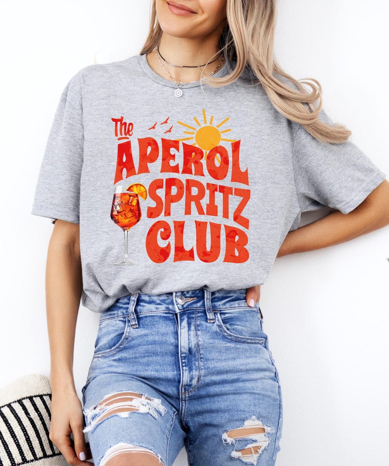K&ouml;nnte beinhalten: Ein graues T-Shirt mit dem Text "The Aperol Spritz Club" in roten und orangen Buchstaben. Das Design enth&auml;lt auch ein Glas Aperol Spritz, eine Sonne und V&ouml;gel.