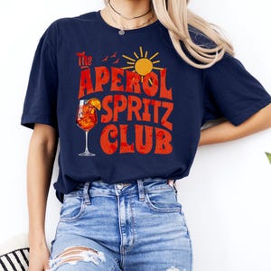K&ouml;nnte beinhalten: Ein marineblaues T-Shirt mit dem Text "The Aperol Spritz Club" in roten und orangen Buchstaben. Das Design enth&auml;lt auch ein Glas Aperol Spritz, eine Sonne und V&ouml;gel.