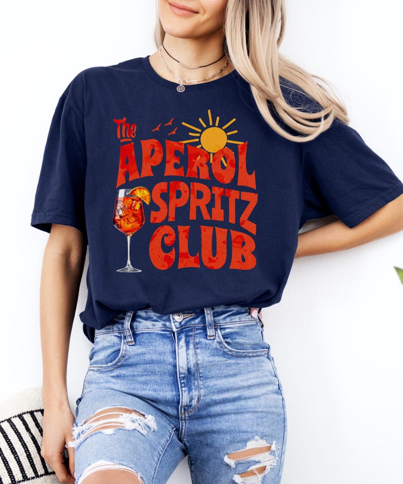 K&ouml;nnte beinhalten: Ein marineblaues T-Shirt mit dem Text "The Aperol Spritz Club" in roten und orangen Buchstaben. Das Design enth&auml;lt auch ein Glas Aperol Spritz, eine Sonne und V&ouml;gel.