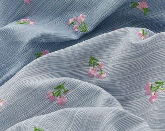 May include: Close-up of a light blue fabric with a subtle striped pattern. The fabric is adorned with delicate embroidered pink flowers and green leaves, creating a soft, textured appearance. The folds in the fabric add depth and dimension to the design.