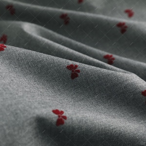 May include: Close-up of grey fabric featuring a repeating pattern of small, red floral designs. The fabric is slightly creased, creating shadows and depth. The floral pattern is evenly spaced across the material.