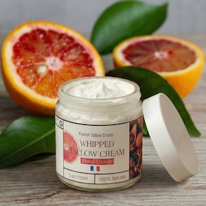 Könnte beinhalten: Ein offenes Glas French Tallow Cream, beschriftet mit "WHIPPED TALLOW CREAM Blood Orange", mit weißer Creme. Der Deckel des Glases liegt daneben. Blutorangenscheiben und grüne Blätter sind im Hintergrund. Das Glas ist 150 ml.