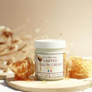 Op de afbeelding: Een pot Franse Talgcrème met de woorden "Whipped Tallow Cream Honey" op het etiket. De pot staat naast stukjes honingraat op een houten oppervlak. Het etiket zegt ook "5 oz | 150 ml" en "100% Natural".