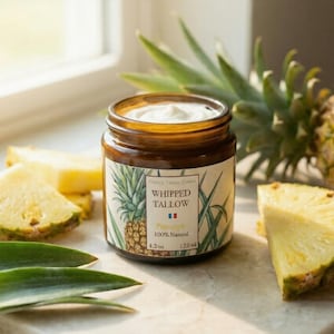 Puede incluir: Un tarro de crema de sebo batida con una etiqueta de piña, rodeado de rodajas de piña fresca y una piña entera. El tarro es de vidrio marrón, y la crema del interior es blanca. La etiqueta dice "Whipped Tallow" y "Pineapple 100% Natural".