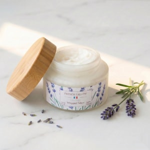 An open glass jar of French Tallow Lavender Whipped Tallow with its bamboo lid leaning against it, sitting on a white marble surface next to sprigs of dried lavender and scattered buds in bright sunlight.