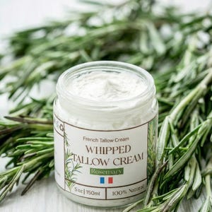 Puede incluir: Un tarro de vidrio transparente lleno de crema batida blanca, etiquetado como "Whipped Tallow Cream Rosemary". El tarro está rodeado de ramitas de romero fresco. La etiqueta indica 150 ml y "100% natural".