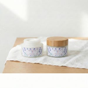 Two frosted glass jars of French Tallow Lavender Whipped Tallow sitting on a white linen cloth over a wooden surface. The jar on the left is open, revealing white cream, while the jar on the right is closed with a bamboo lid.