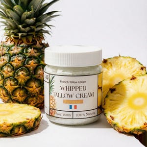 Op de afbeelding: Een pot Franse Tallow Cream met een witte deksel en een etiket met de tekst "Whipped Tallow Cream Pineapple". De pot staat naast een hele ananas en ananasschijfjes. De crème is 150 ml en is 100% natuurlijk.