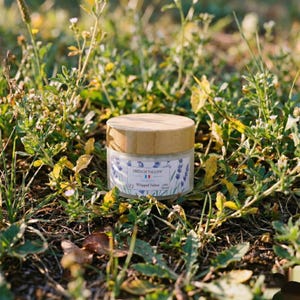 A closed jar of French Tallow Lavender Whipped Tallow sitting directly on the ground, nestled among green grass, leaves, and small wildflowers in natural sunlight.