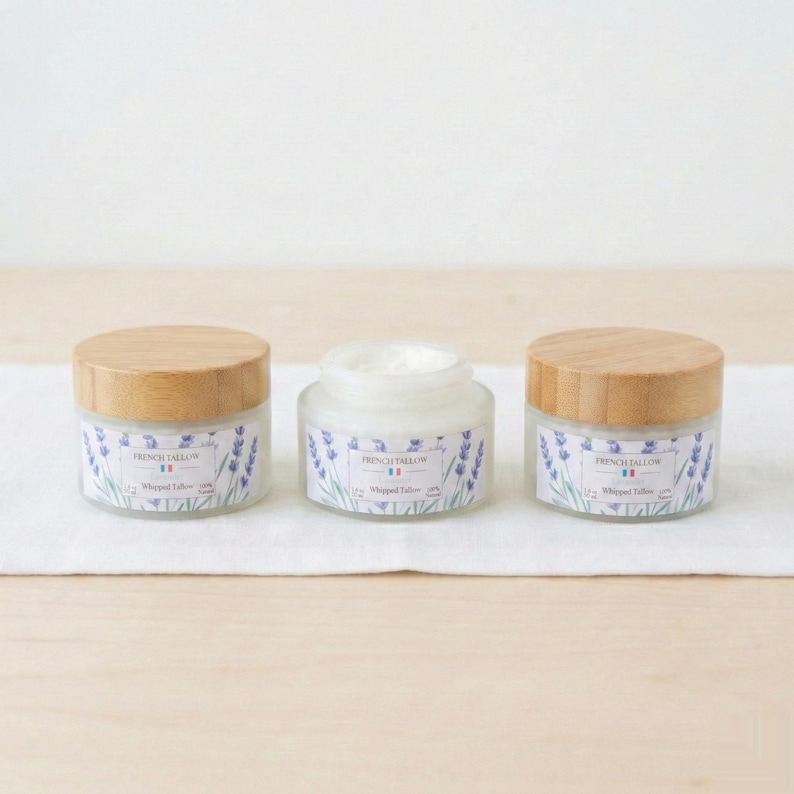 Three jars of French Tallow Lavender Whipped Tallow lined up on a white cloth over a wooden table. The center jar is open, showing the white cream inside, flanked by two closed jars with bamboo lids.