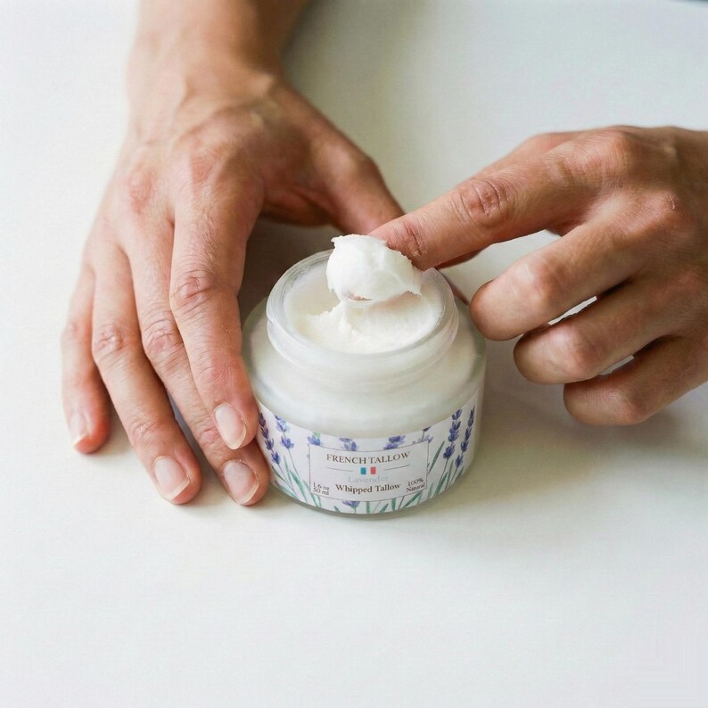 A person's hands holding an open jar of French Tallow Lavender Whipped Tallow, with one finger scooping out a dollop of the thick, white cream against a white background.