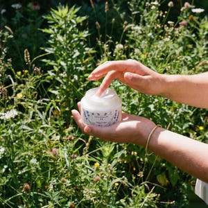 A person's hands holding an open jar of French Tallow Lavender Whipped Tallow outdoors against a background of lush green plants and wildflowers, with one finger scooping out the cream.
