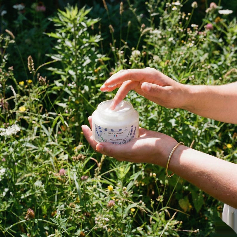 A person's hands holding an open jar of French Tallow Lavender Whipped Tallow outdoors against a background of lush green plants and wildflowers, with one finger scooping out the cream.