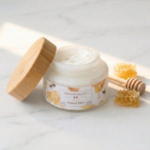 Op de afbeelding: Een glazen pot met opgeklopte talk met een houten deksel, met het label "French Tallow". De pot staat naast stukjes honingraat en een houten honingdipper. Het label heeft een bijengrafiek en de woorden "Whipped Tallow 100% Natural."