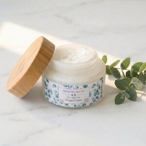 Op de afbeelding: Een open pot "French Tallow Eucalyptus" geslagen talk met een houten deksel. De pot heeft een etiket met een blauw-wit bloemmotief. Het product is crèmewit. Een takje eucalyptus is zichtbaar op de achtergrond.