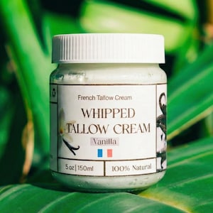 Könnte beinhalten: Ein kleines Glas mit französischem Talgcreme und einem weißen Deckel. Auf dem Etikett steht "WHIPPED TALLOW CREAM Vanilla" mit der französischen Flagge. Das Glas enthält 150 ml Creme, umgeben von grünen Blättern.