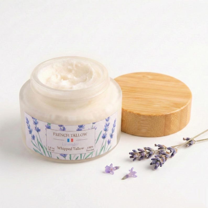 A close-up of an open jar of French Tallow Lavender Whipped Tallow, showing the textured white cream inside. The bamboo lid rests flat next to it alongside sprigs of dried lavender against a plain white background.