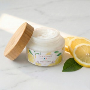 Op de afbeelding: Een pot French Tallow Lemon slagroom met een houten deksel, naast citroenschijfjes en een groen blad. De pot heeft een label met de tekst "French Tallow Lemon Whipped Tallow 100% Natural".