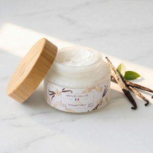 Może przedstawiać: Słoik kremu French Tallow Vanilla z drewnianą pokrywką, obok lasek wanilii i liści. Słoik ma etykietę z napisem "French Tallow Vanilla Whipped Tallow 100% Natural". Słoik zawiera 50 ml.