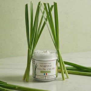 Puede incluir: Un tarro de French Tallow Cream Whipped Tallow Cream con hierba de limón, 150 ml, e ingredientes 100% naturales. El tarro está rodeado de tallos de hierba de limón verde fresco, sobre un fondo verde claro.