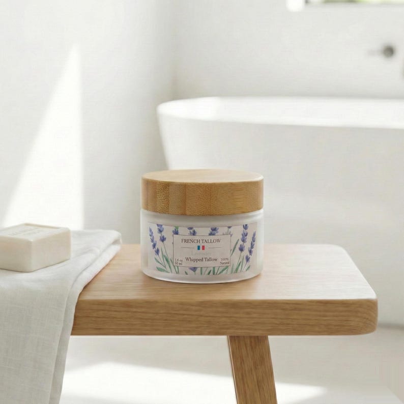 A closed jar of French Tallow Lavender Whipped Tallow sitting on a wooden bath stool next to a folded white towel and a bar of soap, with a blurred white bathtub in the background.