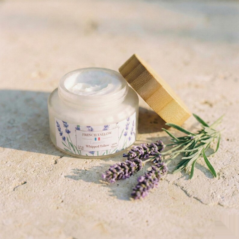 An open jar of French Tallow Lavender Whipped Tallow resting on a textured stone surface outdoors, surrounded by fresh sprigs of lavender, with the bamboo lid leaning nearby.