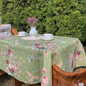 May include: A green tablecloth with a floral pattern featuring pink and red roses, green leaves, and white flowers. The tablecloth is laid on a wicker table with two white teacups and a white vase with pink flowers.