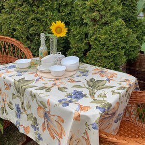 May include: A white tablecloth with a floral pattern featuring orange, blue, and green hues. The tablecloth is laid out on a wicker table with white ceramic cups and saucers, a glass vase with a sunflower, and a glass bottle.