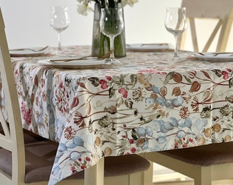 May include: A white tablecloth with a floral pattern in shades of pink, blue, and brown. The tablecloth is laid on a table with two chairs and a vase of white flowers in the center.