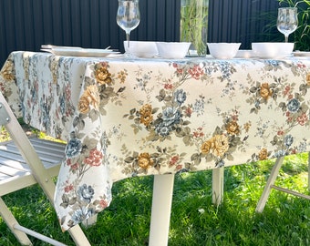 May include: A white table with a floral tablecloth set for a meal outdoors. The table is set with white plates, glasses, and bowls. The table is surrounded by green grass.