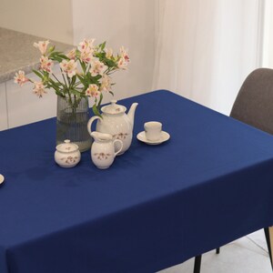 May include: A blue tablecloth with a white teapot, creamer, sugar bowl, and two teacups and saucers. A vase with pink and white flowers is on the table.