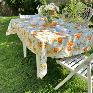 May include: A white tablecloth with a floral pattern featuring orange fruit and yellow flowers. The tablecloth is laid out on a table with white chairs and a bouquet of white flowers in a glass vase.