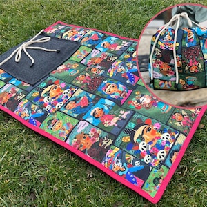 Op de afbeelding: Een kleurrijk picknickkleed met een zwarte zak en een roze rand. Het kleed heeft een herhalend patroon van kleurrijke illustraties van mensen met bloemen en schedels. Een bijpassende trekkoordtas is ook afgebeeld.