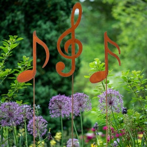 Può includere: Tre paletti da giardino in metallo a forma di note musicali. I paletti sono di colore marrone ruggine e sono posizionati in un giardino con fiori viola.