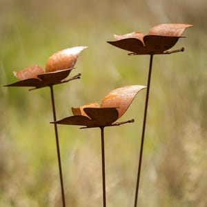 Op de afbeelding: Drie roestige metalen vlinders op dunne metalen stelen. De vlinders zijn bruin en hebben een licht getextureerd oppervlak.