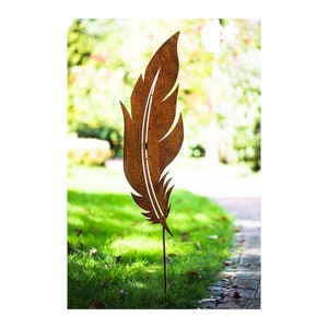 Op de afbeelding: Een roestige metalen tuinpaal in de vorm van een veer. De paal staat in een grasachtig gebied met een bakstenen pad op de achtergrond.