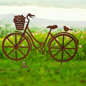 Op de afbeelding: Een roestige metalen silhouet van een fiets met een mand vol bloemen en een vogel die op het achterwiel zit. De fiets staat in een grasveld.