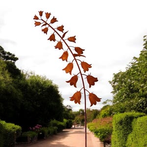 Op de afbeelding: Een hoog, roestig metalen beeld van een bloem met klokvormige bloesems. Het beeld staat in een tuin met groene bomen en struiken op de achtergrond.