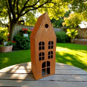 Op de afbeelding: Een roestbruin metalen huisvormig tuinornament. Het huis heeft uitgesneden ramen en een deur, met een cirkelvormige opening nabij de top van het dak. Het metaal heeft een verweerde uitstraling. Het huis staat op een houten tafel.