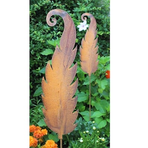 Könnte beinhalten: Zwei rostige Metall-Gartenstecker in Form von stilisierten Blättern mit einer gekräuselten Spitze. Die Stecker befinden sich in einer Gartenumgebung mit grünem Laub und Blumen.