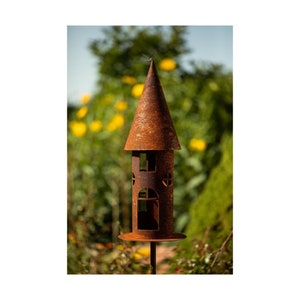 Op de afbeelding: Een roestig metalen vogelhuisje in de vorm van een toren met een puntdak. Het vogelhuisje heeft verschillende uitgesneden ramen en is gemonteerd op een metalen paal.