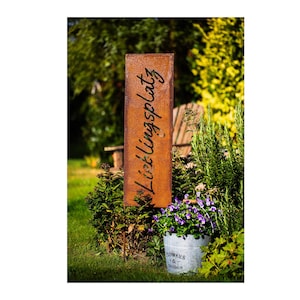 Może przedstawiać: Rustykalny metalowy znak z napisem "Lieblingsplatz" w ozdobnym, pochylonym piśmie. Znak stoi w ogrodzie, na tle zielonej trawy i doniczki z fioletowymi kwiatami.